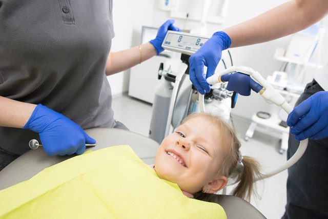 Smiling young girl with ponytail reclined in a dentist's chair with two blue gloved dental professionals.
